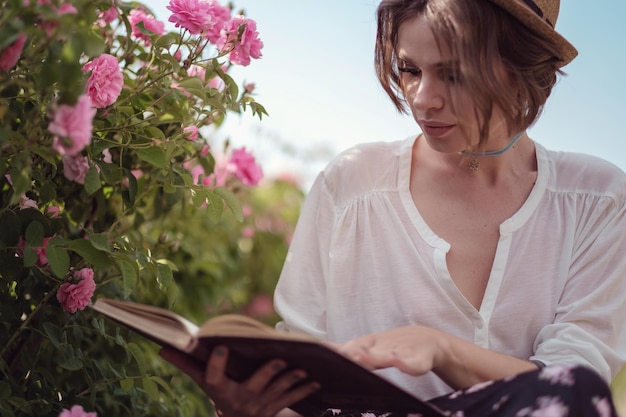 Mooi meisje met hoed met boek zittend op gras in rose gaden een plattelandsmeisje geniet van de zonsondergangwijn en leest boeken in de geurige rozentuin het concept van parfum Premium Foto