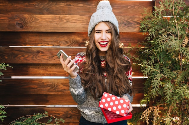 Mooi meisje met lang haar en rode lippen met kerstdoos en telefoon op houten. Ze draagt warme winterkleren, glimlachend.