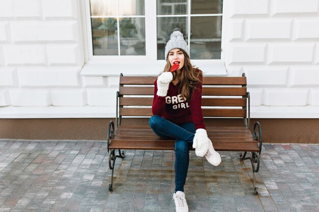 Mooi meisje met lang haar in gebreide mutsjeans, witte handschoenen, zittend op een bankje op straat. Ze kijkt genoten naar het likken van karamelhart.