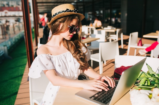 Mooi meisje met lang haar in hoed zit aan tafel op het terras in café. Ze draagt een witte jurk met blote schouders, rode lippenstift, zonnebril. Ze typt serieus op laptop.