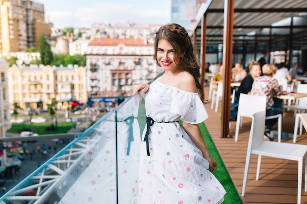 Mooi meisje met lang haar is poseren voor de camera op het terras in café. Ze draagt een witte jurk met blote schouders en rode lippenstift.