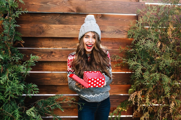 Mooi meisje met lang haar met kerstdoos op houten. ze draagt warme winterkleren, een gebreide muts en glimlacht blij.