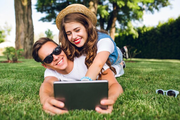 Mooi meisje met lang krullend haar en rode lippen ligt op de achterkant van een knappe jongen in een zonnebril op gras in het zomerpark. Guy houdt tablet vast en ze hebben plezier.