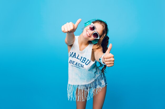 Mooi meisje met lang krullend haar in staart in blauwe zonnebril op blauwe achtergrond in de studio. Ze draagt een wit T-shirt, korte broek en luistert naar muziek met een blauwe koptelefoon, ziet er genoten en gelukkig uit.