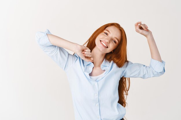 Mooi meisje met lang rood haar vingers knippen en dansen, genieten van muziek luisteren, staande tegen een witte muur