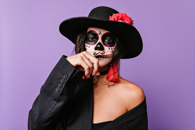 Mooi meisje met ongebruikelijke gezichtskunst in Mexicaanse stijl, die haar vinger met geschilderde snor voor haar lippen houdt.