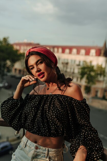 Mooi meisje met rode lippen en zilveren accessoires in polka dot wijde blouse met lange mouwen en lichte spijkerbroek op zoek naar camera op balkon