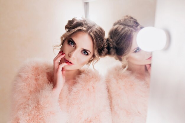 Mooi meisje met schattig kapsel aanraken van haar gezicht tijdens het wachten op fotoshoot in de kleedkamer. Schitterende gekrulde jonge vrouw in roze jasje op zoek met interesse poseren naast make-up spiegel