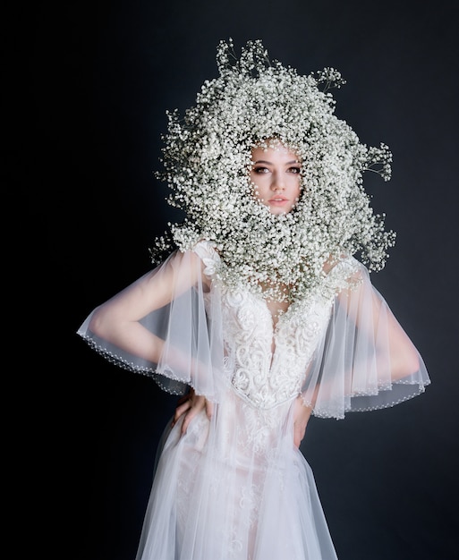 Mooi meisje met verse gypsophila krans op het gezicht gekleed in witte tedere jurk op de donkere achtergrond