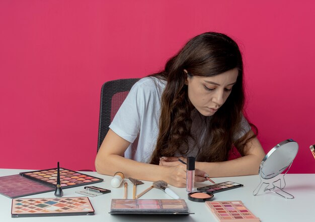 Mooi meisje zit aan de make-up tafel met make-up tools oogschaduw borstel te houden en te kijken naar oogschaduw palet geïsoleerd op crimson achtergrond