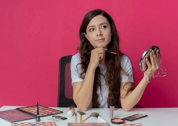 Mooi meisje zit aan de make-up tafel met make-up tools spiegel en oogschaduw borstel te houden kant kijken en kin geïsoleerd op crimson achtergrond aan te raken