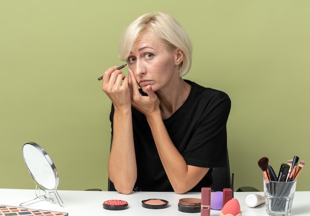 Gratis foto mooi meisje zit aan tafel met make-up tools teken pijl met eyeliner geïsoleerd op olijf groene achtergrond