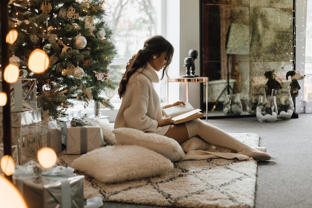 Mooi meisje zit met kussens en plaid in de buurt van een kerstboom en een boek te lezen