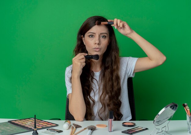 Mooi meisje, zittend aan de make-up tafel met make-up tools make-up borstels te houden in de buurt van gezicht kijken camera geïsoleerd op groene achtergrond