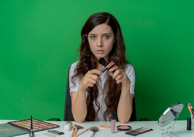 Mooi meisje zittend aan de make-up tafel met make-up tools poeder en blozen borstels te houden en nee gebaren en kijken naar camera geïsoleerd op groene achtergrond