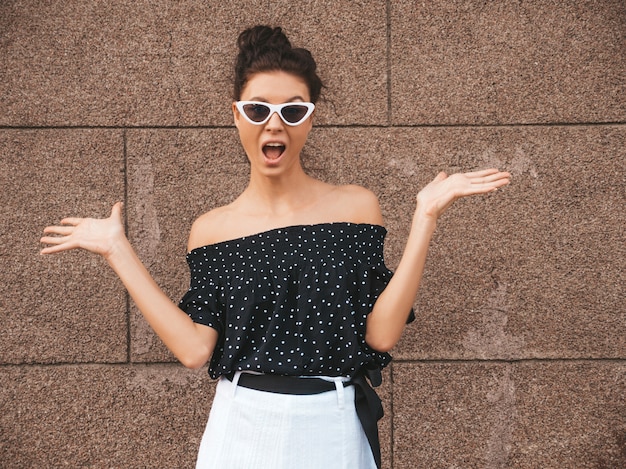 Mooi model gekleed in elegante zomerkleding. Sexy zorgeloos meisje poseren in de straat in de buurt van muur. Trendy moderne zakenvrouw in zonnebril. Iets weergeven op beide vlakke handen voor vergelijkbare keuze