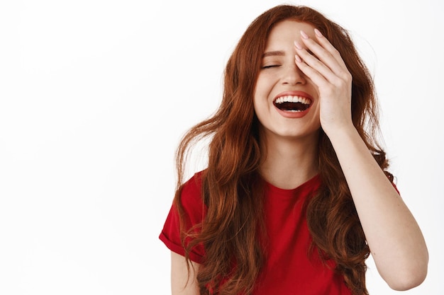 Mooi natuurlijk meisje met gember krullend, oprecht lachen en lachende witte tanden, die de helft van het gezicht bedekken, staande in rood t-shirt tegen witte achtergrond