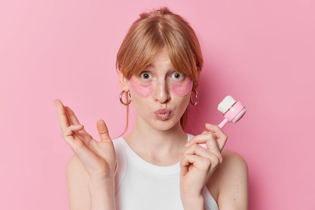 Mooi roodharig meisje houdt de lippen gevouwen brengt hydrogelpleisters onder de ogen aan Gebruikt gezichtsborstel voor massage draagt een casual wit t-shirt geïsoleerd over roze achtergrond Gezichtsverzorging en schoonheidsprocedures
