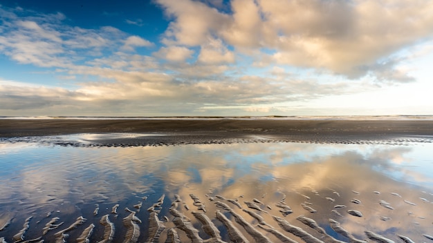Gratis foto mooi schot van een natte zandige kust met watervijver onder een blauwe bewolkte hemel
