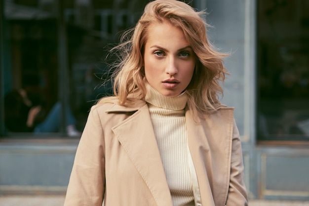 Gratis foto mooi serieus blond meisje in trenchcoat die vol vertrouwen in de camera kijkt op straat in de stad