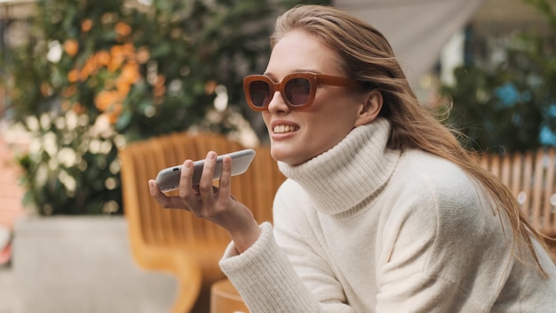 Mooi stijlvol meisje in zonnebril gekleed in een gezellige witte trui die een spraakbericht stuurt naar de beste vriend met een smartphone in het straatcafé. modern technologisch concept