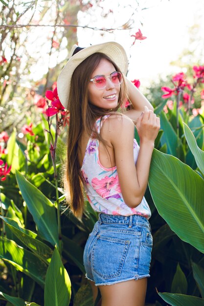 Mooi strandmeisje dat zich in een park bevindt dat strohoed, roze brillen, denimborrels en lichte t-shirt draagt. Het meisje is erg slank en aantrekkelijk. Ze ziet er speels uit. Ze staat tussen tropische bloemen