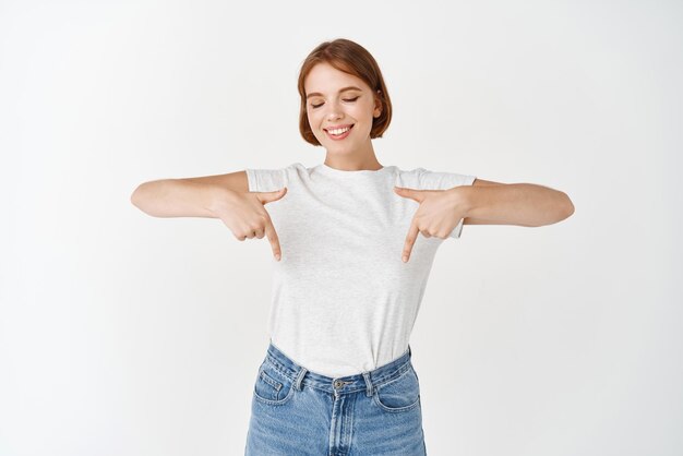 Mooi teder meisje met kort kapsel met een t-shirt dat met de vingers naar beneden wijst en met een glimlach naar de witte achtergrond van de advertentie kijkt