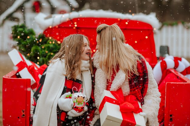 Mooi tienermeisje en blonde aantrekkelijke moeder in witte en rode winterkleren die in de kerstauto zitten en lachen onder het sneeuwen.