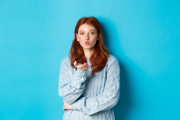 Mooi tienermeisje in trui die luchtkus blaast, lippen tuit en naar de camera staart, staande tegen een blauwe achtergrond