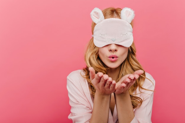 Mooi verlegen meisje met blonde haren poseren in slaapmasker. binnen schot van schattig kaukasisch vrouwelijk model verzendt luchtkus in oogmasker.