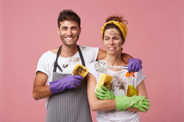 Mooi verliefd stel dat klusjes doet: aantrekkelijke man die zijn vrouw omhelst die met afkeer naar vuile borstel kijkt. Teamwerk van familie die aan huis werkt. Knappe man en vrouw wassen van meubels