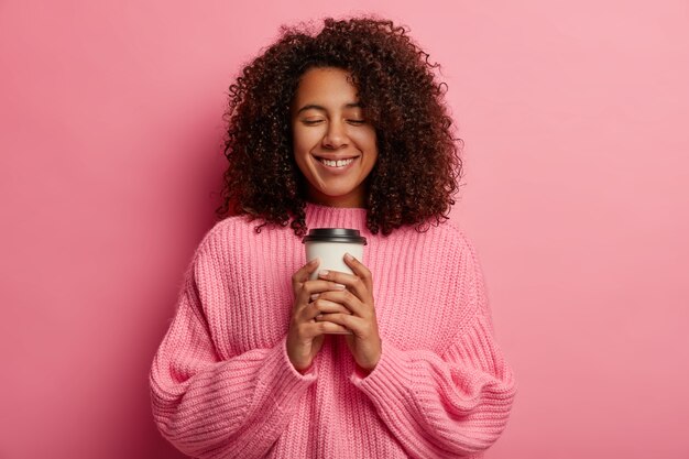 Mooi vrolijk duizendjarig meisje met zachte glimlach, houdt afhaalkoffie vast, geniet van aromatische cafeïnedrank en goede smaak