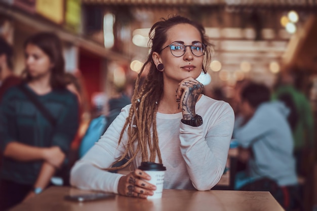 Mooi vrolijk meisje met tatoeages en dreadlocks zit aan de food court terwijl ze koffie drinkt.