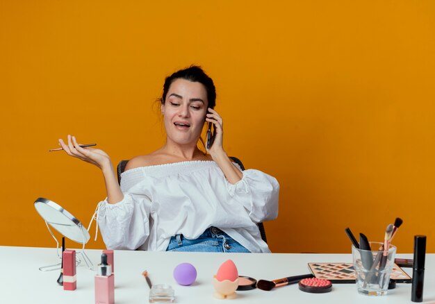 Mooi vrolijk meisje zit aan tafel met make-up tools gesprekken over telefoon met make-up borstel geïsoleerd op oranje muur