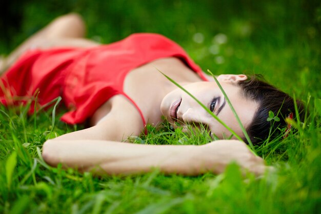 mooi vrouwelijk donkerbruin meisjesmodel dat in het groene heldere gras van de zomer in het park met make-up in rode kleding ligt