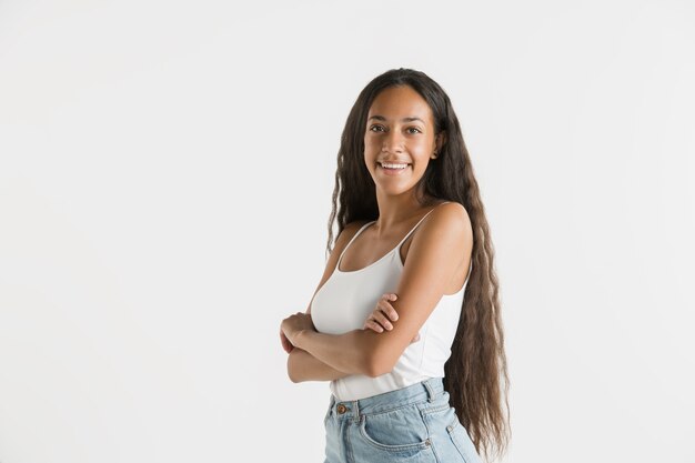 Mooi vrouwelijk half-lengteportret dat op witte muur wordt geïsoleerd. Jonge emotionele Afro-Amerikaanse vrouw met lang haar. Gelaatsuitdrukking, concept van menselijke emoties. Staan en glimlachen.