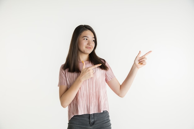 Mooi vrouwelijk half-lengteportret dat op witte studioachtergrond wordt geïsoleerd. Jonge emotionele vrouw in vrijetijdskleding. Menselijke emoties, gezichtsuitdrukking concept. Naar opzij wijzend, glimlachend.