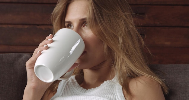 Mooi vrouwelijk model op een bank met een kopje koffie onder een blauwe deken, kijkend naar de camera en teruggaand naar haar gedachten