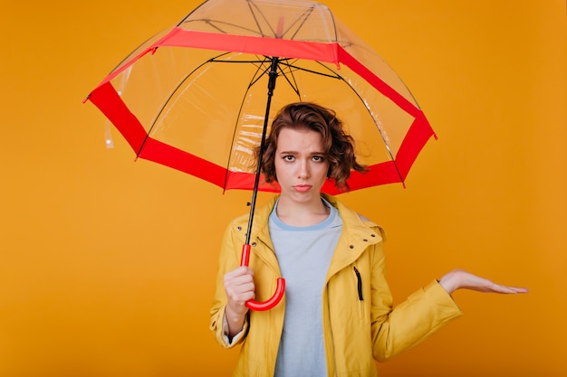 Mooi wit meisje droevige emoties uitdrukken terwijl ze onder de paraplu staat. Binnenfoto van verlegen, overstuur dame draagt herfstkleding met parasol.