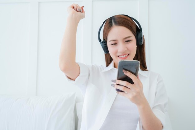 Mooie aantrekkelijke aziatische vrouw luistert lied van hoofdtelefoon hand houdt smartphone witte slaapkamer achtergrond portret van aziatische lang haar wit overhemd vrouw