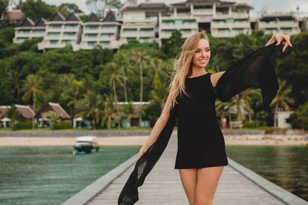 Mooie aantrekkelijke vrouw gekleed in zwarte jurk poseren op pier in luxeresort, zomervakantie, tropisch strand