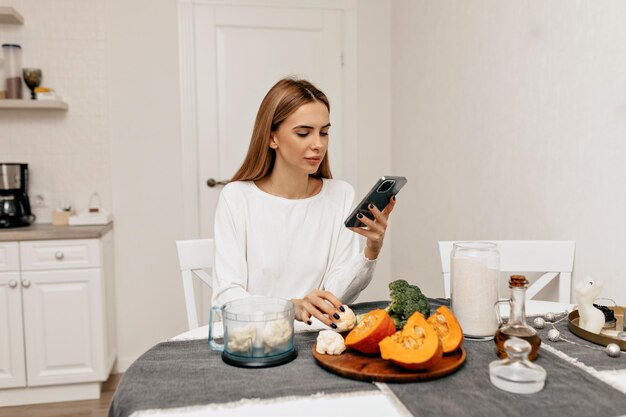 Mooie aantrekkelijke vrouw met donker haar die een wit overhemd draagt, kijkt in het recept van een smartphone Vrouw in de keuken met verse groenten aan het koken