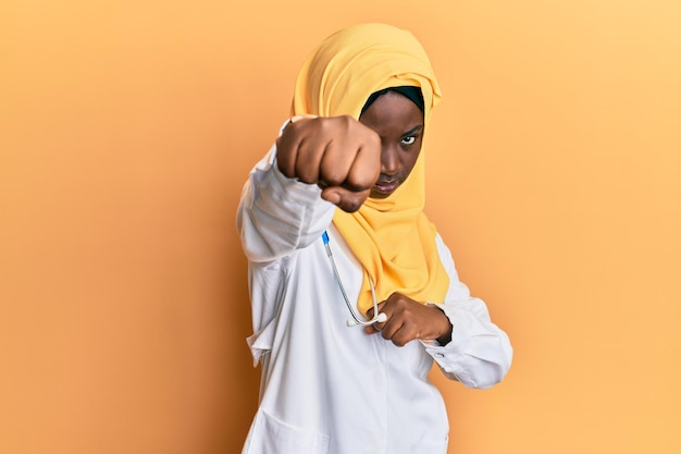Gratis foto mooie afrikaanse jonge vrouw in doktersuniform en hijab ponsvuist om te vechten, agressieve en boze aanval, dreiging en geweld