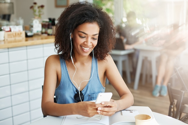 Mooie Afrikaanse vrouw in hoofdtelefoons glimlachen die de zitting van het telefoonscherm bekijken die in koffie rusten.