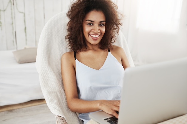 Mooie Afrikaanse vrouw in nachtkleding glimlachend zittend in stoel met laptop thuis.