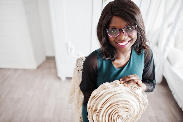 Mooie Afro-Amerikaanse vrouw in brillen poseerde in de kamer zittend op een stoel met decoratieve bloem in handen