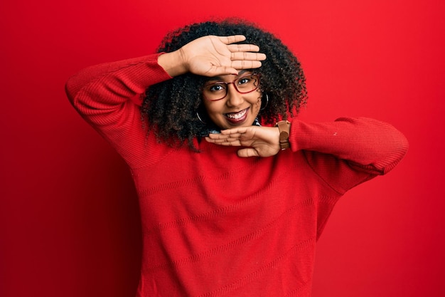 Mooie afro-amerikaanse vrouw met afrohaar in trui en bril glimlachend vrolijk spelen peek a boo met handen die gezicht laten zien. verrast en uitgelaten