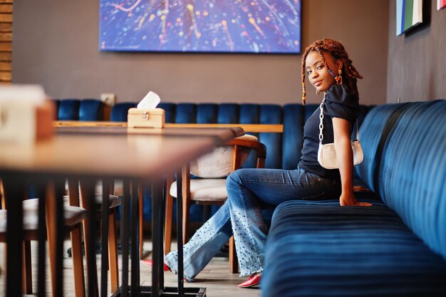 Mooie Afro-Amerikaanse vrouw met dreadlocks in blauwe stijlvolle jeans in café Mooi cool, modieus zwart jong meisje binnen