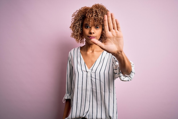 Gratis foto mooie afro-amerikaanse vrouw met krullend haar die een gestreept t-shirt over een roze achtergrond draagt terwijl ze stopt met zingen met de palm van de hand waarschuwingsuitdrukking met een negatief en serieus gebaar op het gezicht