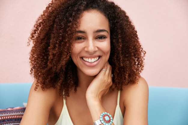 Mooie afro-amerikaanse vrouw met krullend haar, heeft witte perfecte tanden, recreëert binnenshuis tegen roze achtergrond